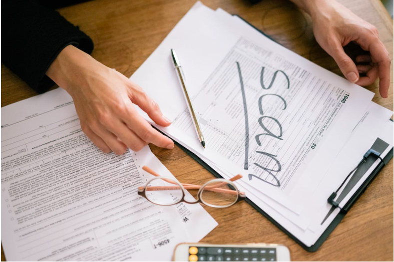 Image shows a Close-Up Shot of a Person Holding Documents with 'scam' written across the front.